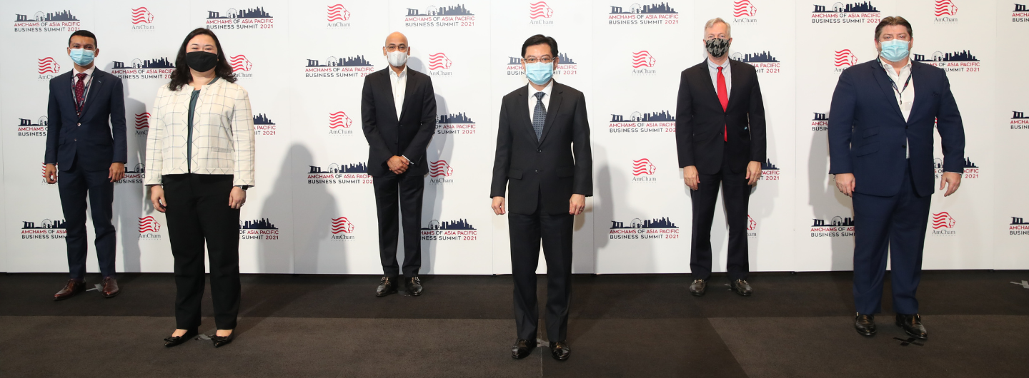 Six people wearing masks and business attire pose in front of an AmCham backdrop.